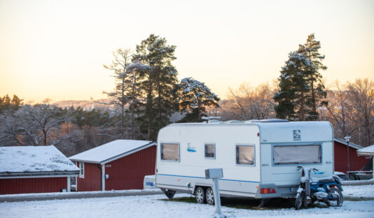 Vinterbild på en husvagn som står på campingplatsen, i bakgrunden syns röda stugor.