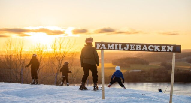 Bild från toppen av Flottsbro skidbacke i solnedgången, skidåkare syns sedda bakifrån, i förgrundens syns en skylt med texten familjebacken.