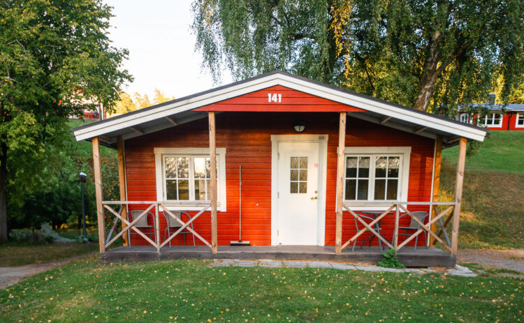 Bild på röd stuga sedd rakt framifrån, gräsmattan är grön och två björkar står på dess vardera sida.